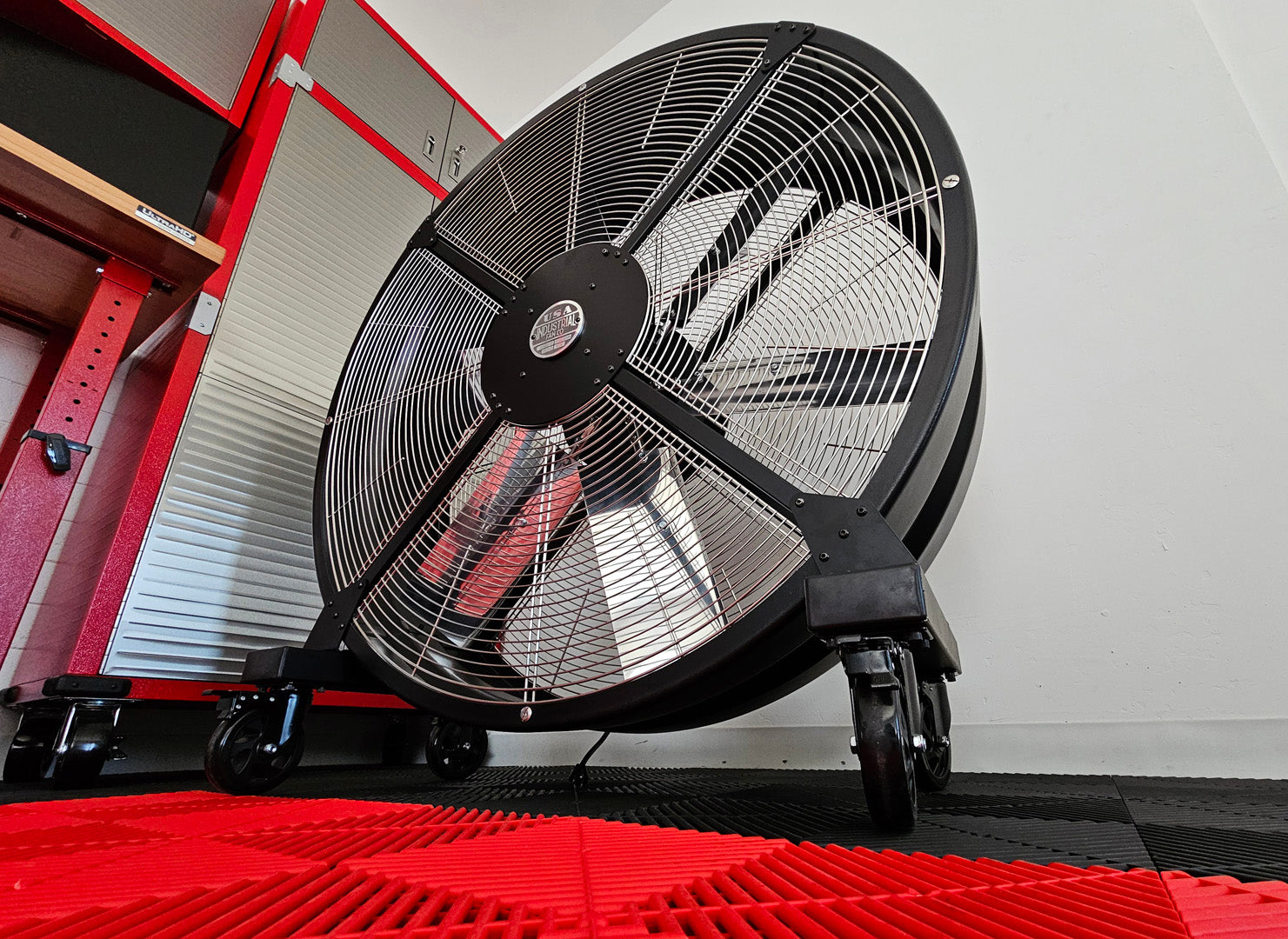 48" industrial floor fan, with black housing, silver blades and silver brand badge, product of Industrial Fan Co, on a red and black floor with a white wall background