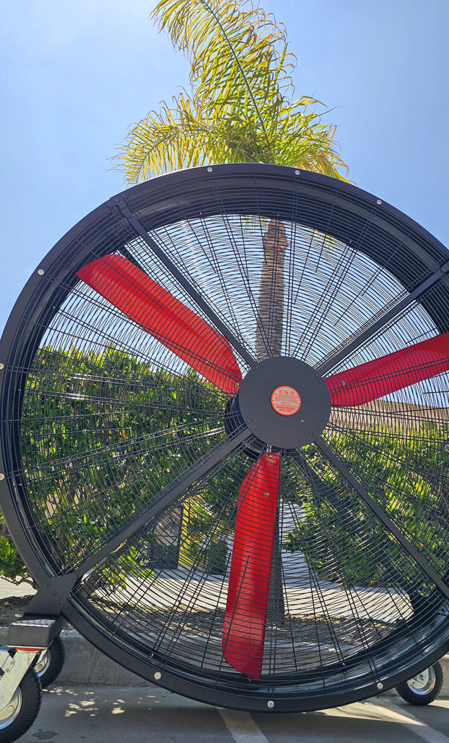 72" Floor Fan with black housing, red blades, metal frame, black and silver casters and metal bearings, product of Industrial Fan Co, with blue sky in the background and palm tree and green bushes