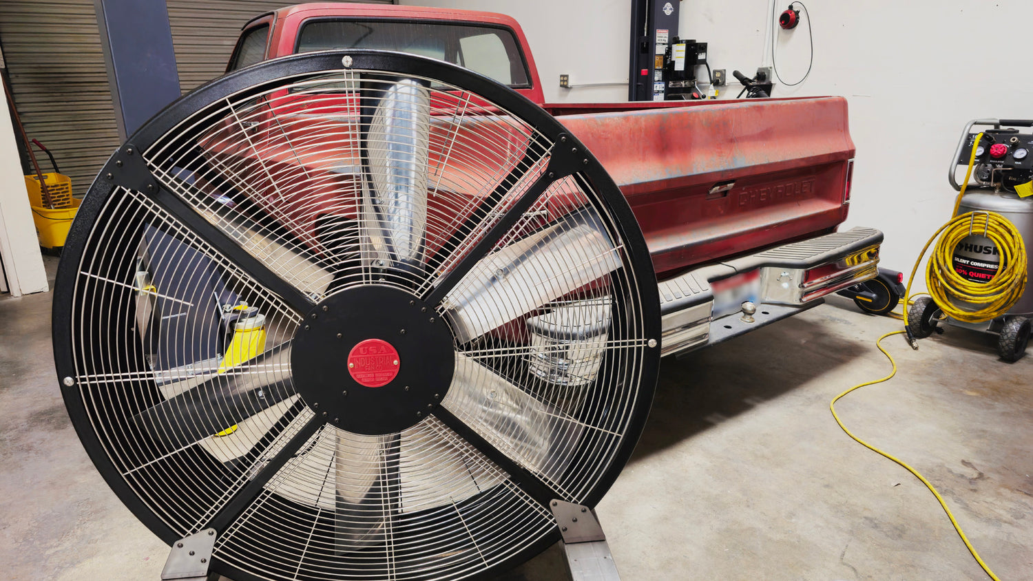 48" industrial floor fan, with black housing, silver blades and red brand badge, product of Industrial Fan Co, with a garage setting background and a red truck