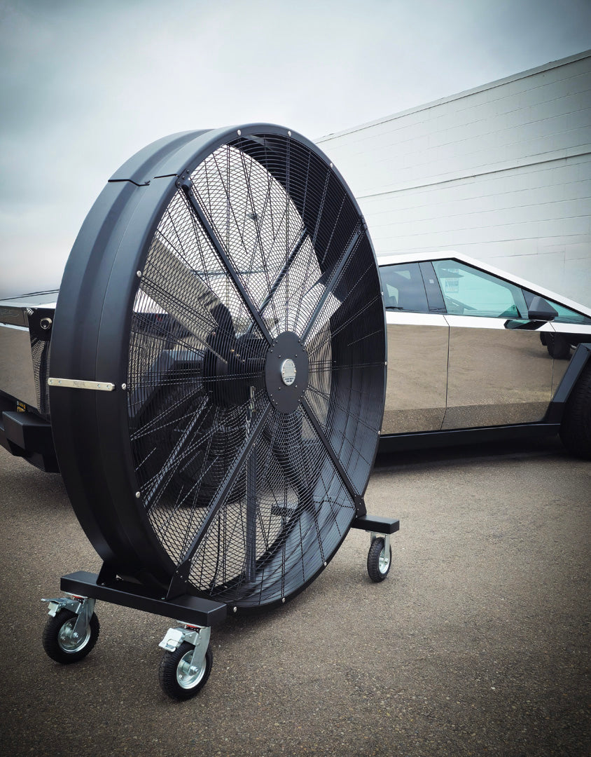 72" Floor Fan with black housing, polished aluminum silver blades, metal frame, black and silver casters and metal bearings, product of Industrial Fan Co, on a parking lot next to a polished silver Tesla CyberTruck
