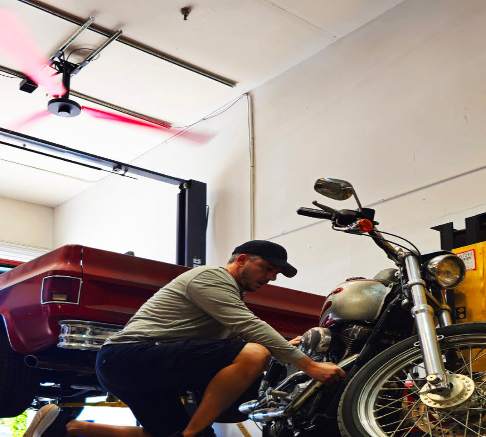 Man working on a motorcycle in a garage with a car in the background and a DF3 Industrial Ceiling Fan above his head, product of Industrial Fan Co