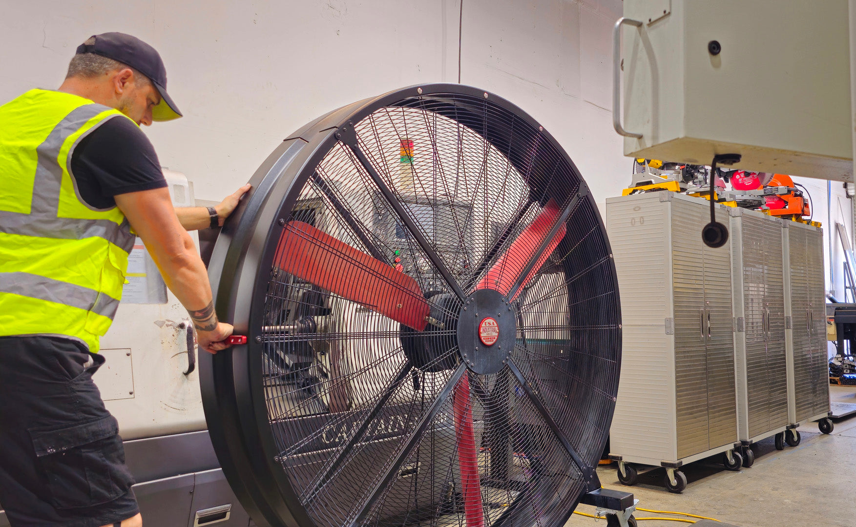 WF72 Industrial Floor Fan, product of Industrial Fan Co, with black housing, red blades, metal frame and red and silver brand badge, being pushed around by a worker in yellow-green vest