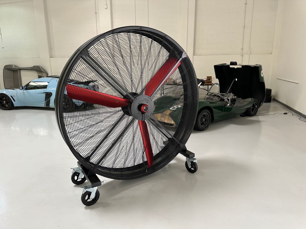 72" Floor Fan with black housing, red blades, metal frame, black and silver casters and metal bearings, product of Industrial Fan Co, inside of a garage with a blue and green sport cars in the background
