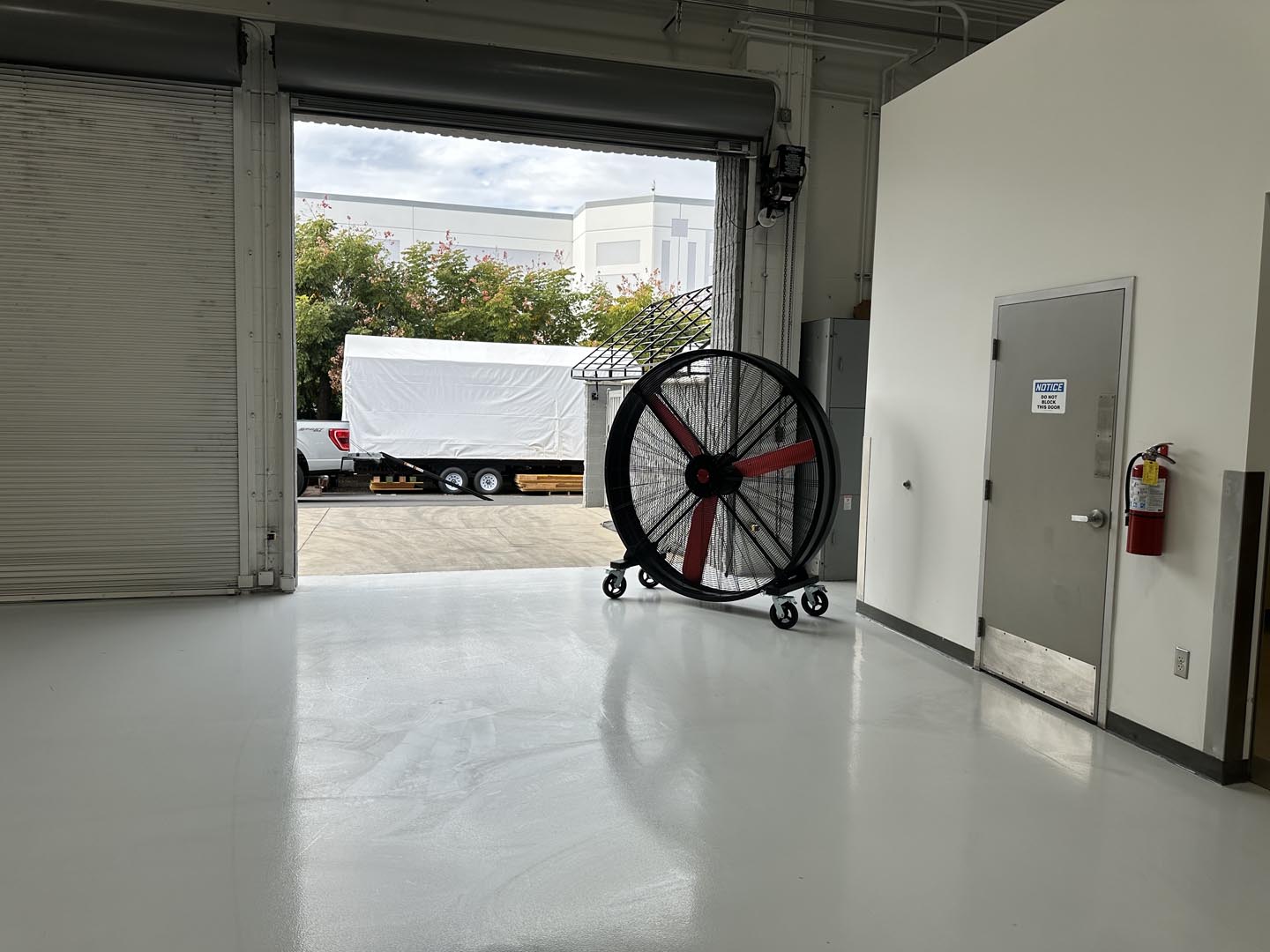 72" Floor Fan with black housing, red blades, metal frame, black and silver casters and metal bearings, product of Industrial Fan Co, in front of a an open garage door in industrial setting