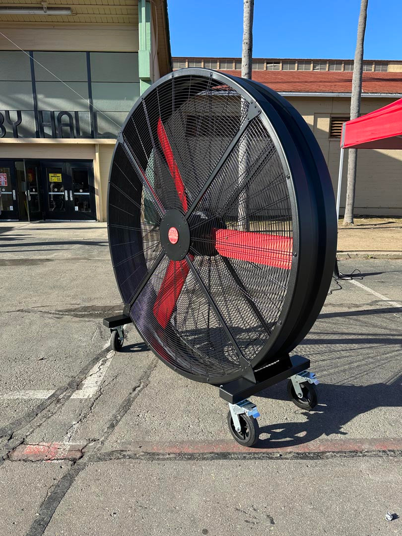 72" Floor Fan with black housing, red blades, metal frame, black and silver casters and metal bearings, product of Industrial Fan Co, in a parking lot with a commercial building in the background