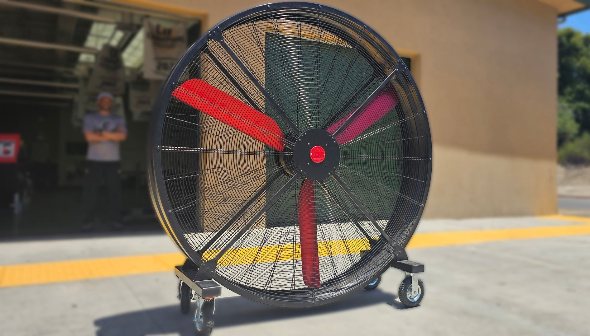 72" Floor Fan with black housing, red blades, metal frame, black and silver casters and metal bearings, product of Industrial Fan Co, in front of a open garage door