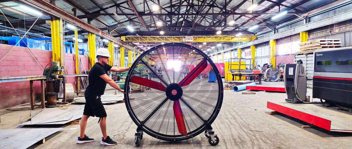 72" Floor Fan with black housing, red blades, metal frame, black and silver casters and metal bearings, product of Industrial Fan Co, in a large industrial hangar being pushed by a man 