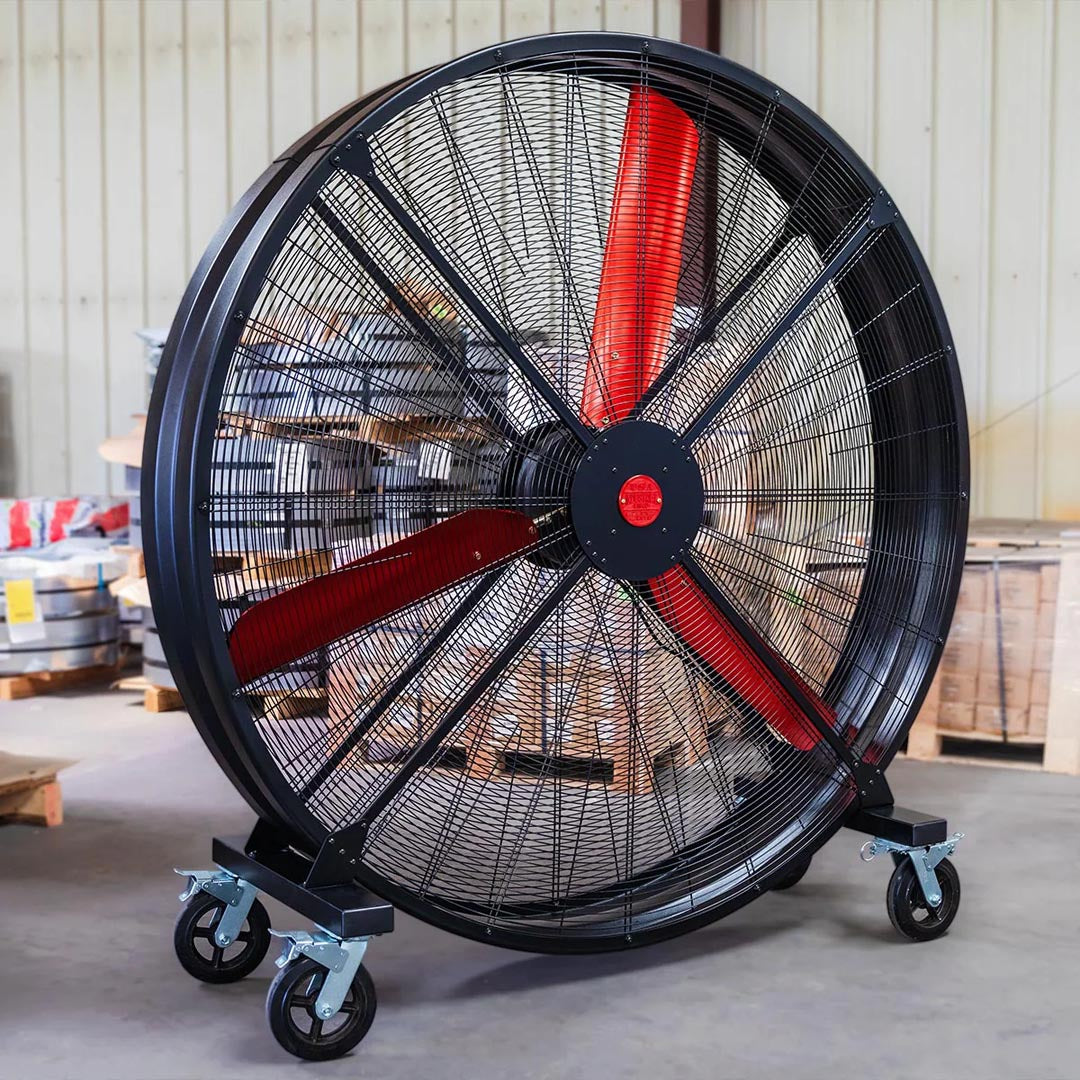 72" Floor Fan with black housing, red blades, metal frame, black and silver casters and metal bearings, product of Industrial Fan Co, inside of a industrial hangar with boxes and industrial material in the background