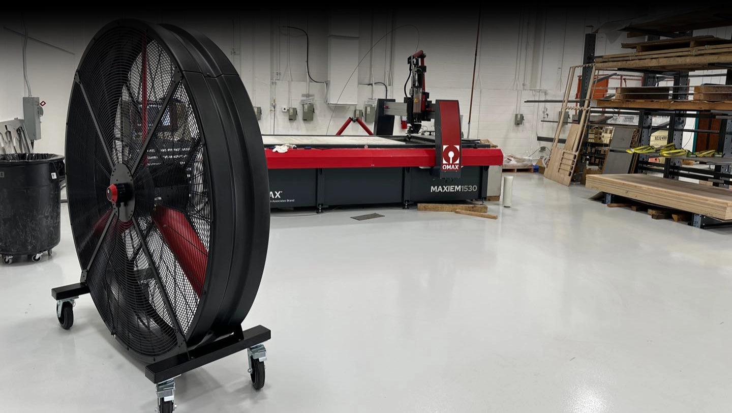 72" Floor Fan with black housing, red blades, metal frame, black and silver casters and metal bearings, product of Industrial Fan Co, inside of a industrial hangar with other industrial machines and stacked wooden palettes in the background.
