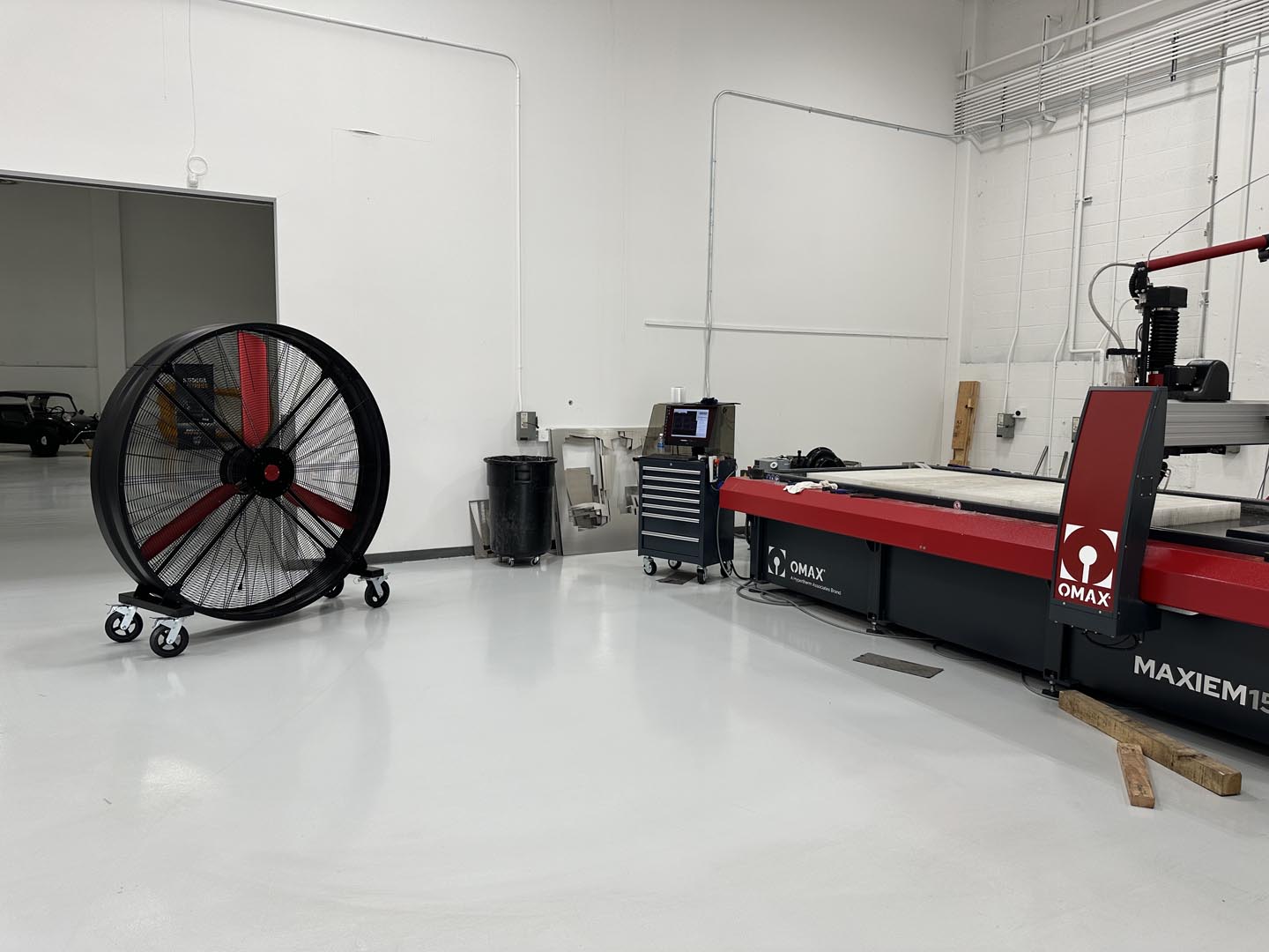 72" Floor Fan with black housing, red blades, metal frame, black and silver casters and metal bearings, product of Industrial Fan Co, inside of a industrial hangar with other industrial machines and a small car deep in the background