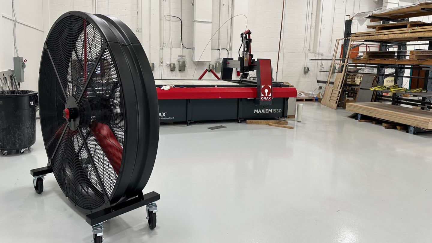 72" Floor Fan with black housing, red blades, metal frame, black and silver casters and metal bearings, product of Industrial Fan Co, inside of a industrial hangar with other industrial machines and stacked wooden palettes in the background