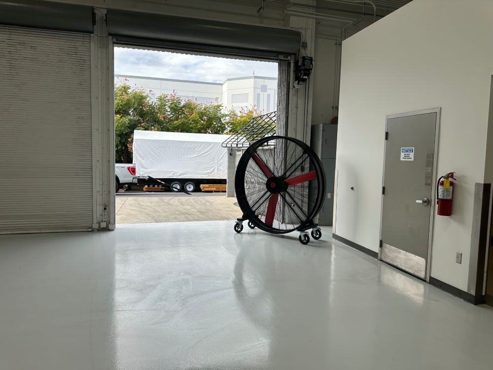 72" Floor Fan with black housing, red blades, metal frame, black and silver casters and metal bearings, product of Industrial Fan Co, in front of a an open garage door in industrial setting